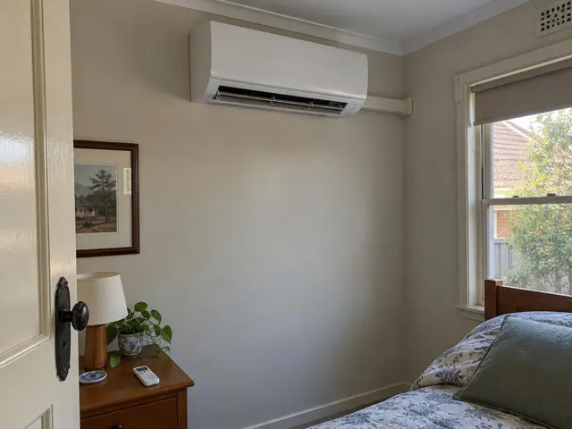 Clean split-system indoor unit high on a bedroom wall with open louvre and a window beside it in Kilburn